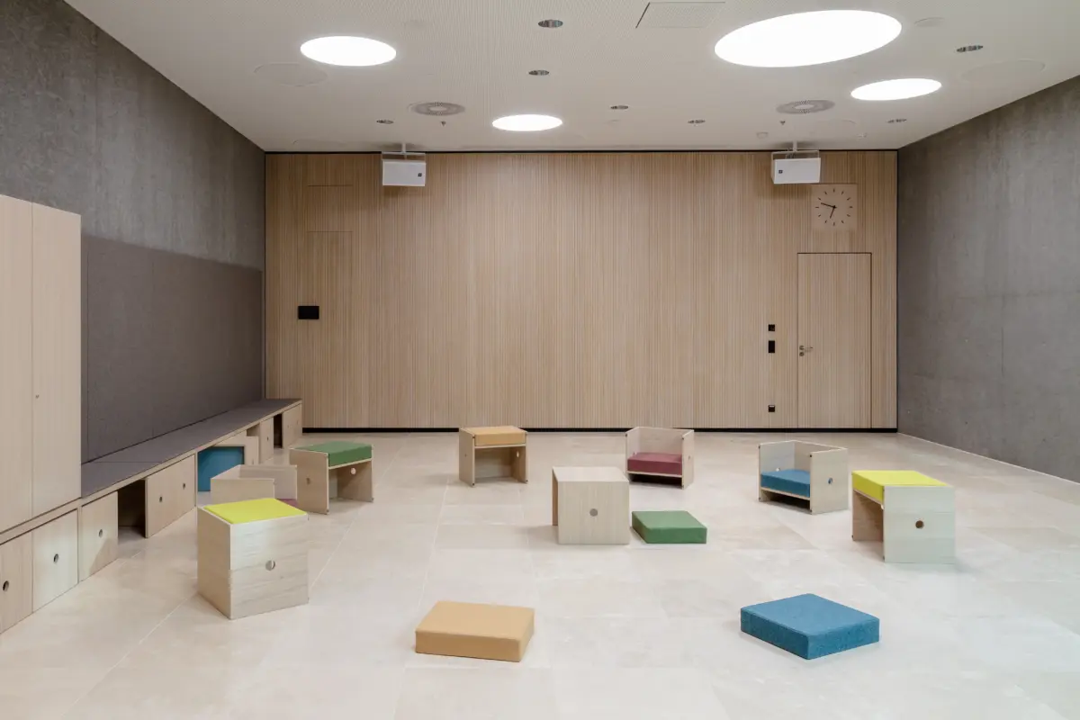 Heller Raum mit holzfarbener Schrankwand, seitlicher Sitzbank und verteilten Sitzwürfeln mit bunten Polstern.