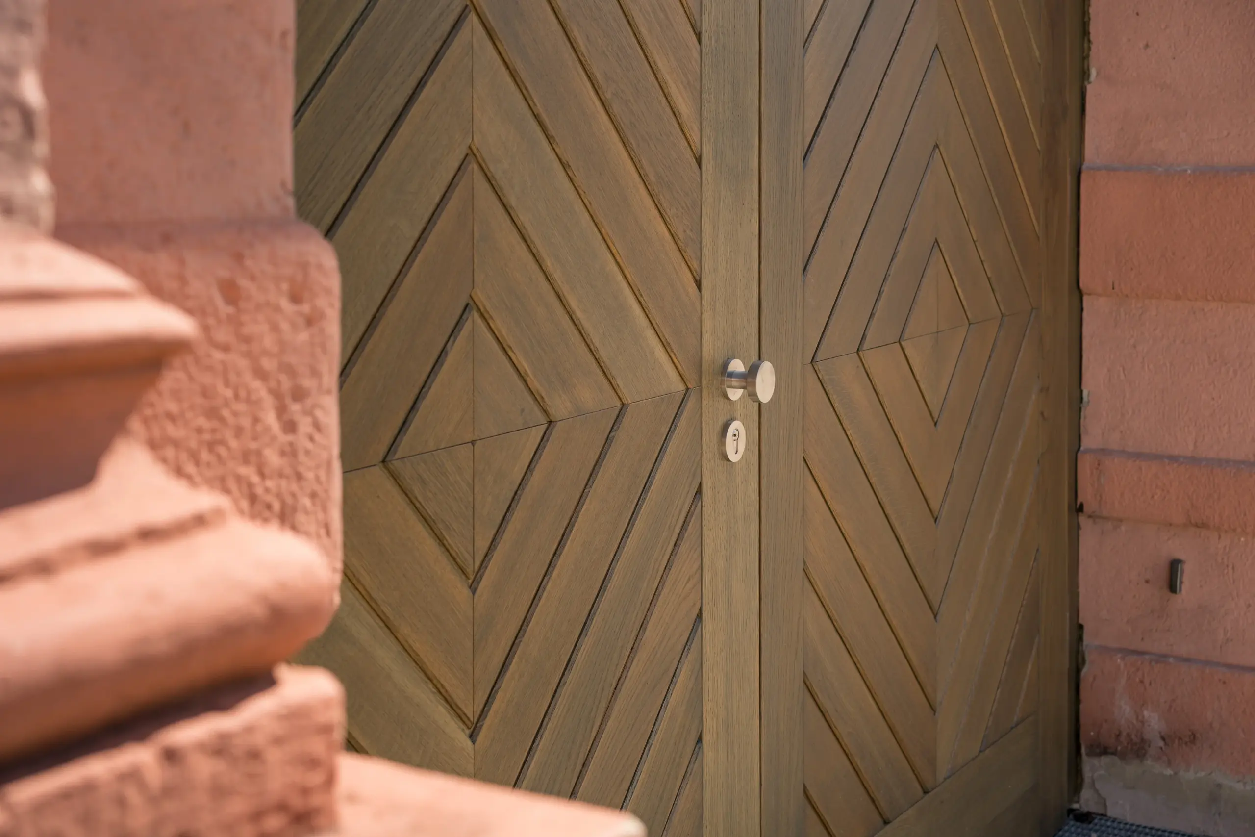 Detail eines Holztores mit rautenförmig angeordneten Brettern, rundem Metallknauf und rötlicher Steineinfassung.