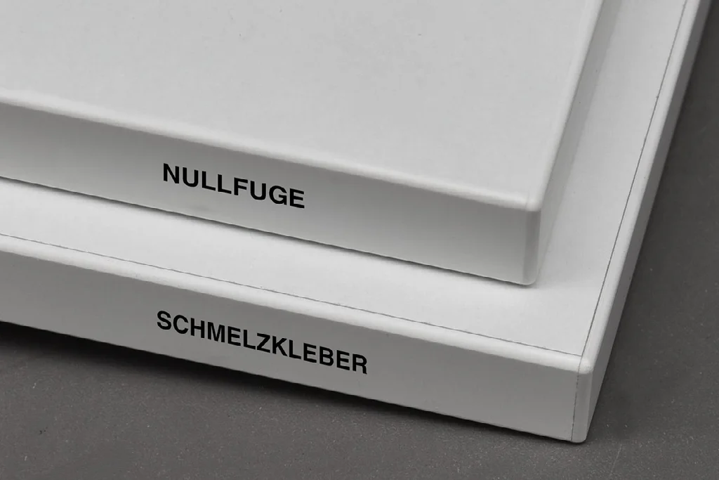 Kantenvergleich: Nullfuge und Schmelzkleber Zwei weiße Plattenkanten im Vergleich, oben mit der Beschriftung Nullfuge und unten mit Schmelzkleber.