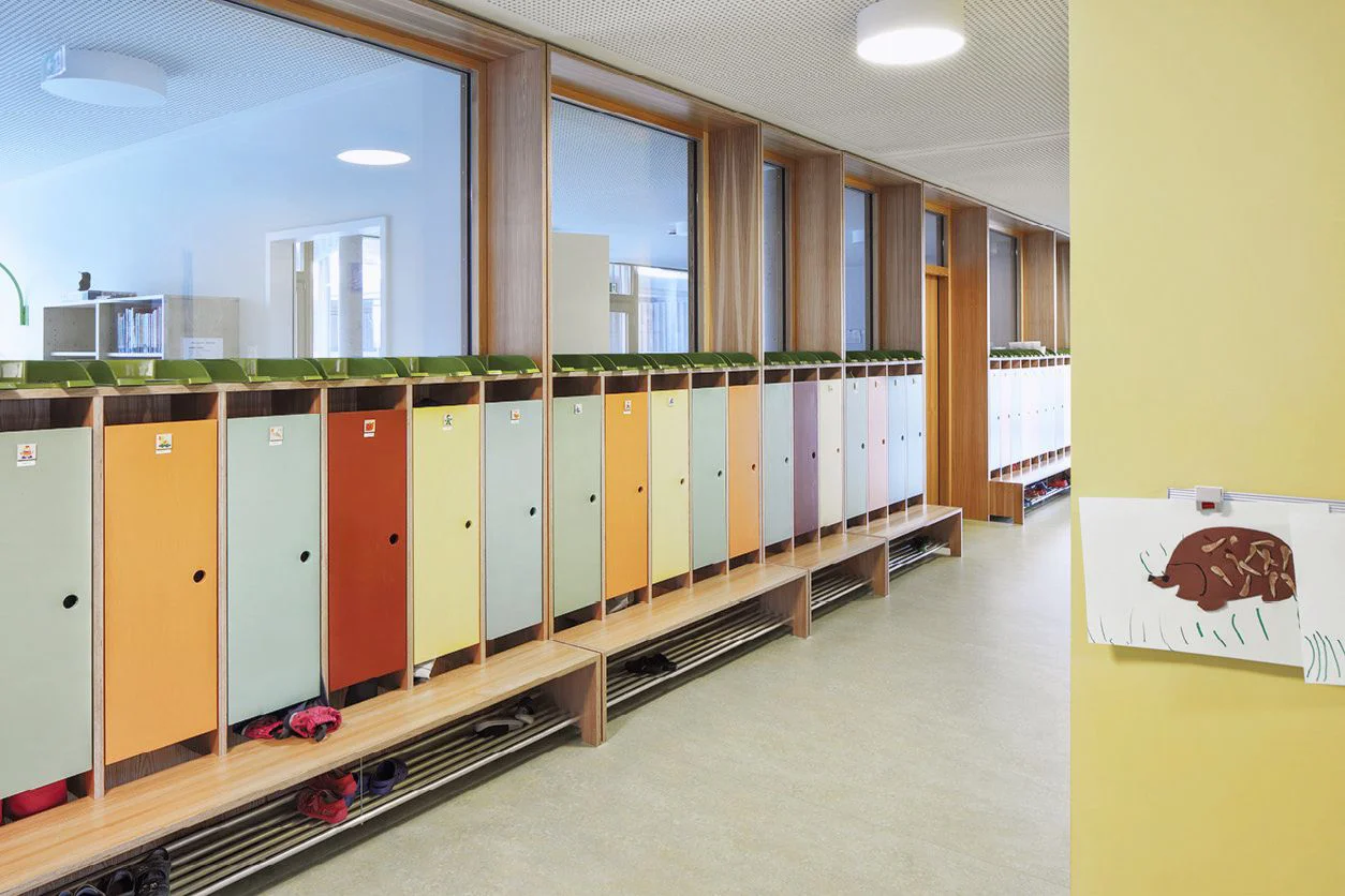 Lange Kindergarderobe mit bunten Schrankfronten, durchgehender Holzsitzbank und offenen Schuhablagen unter großen Fenstern.