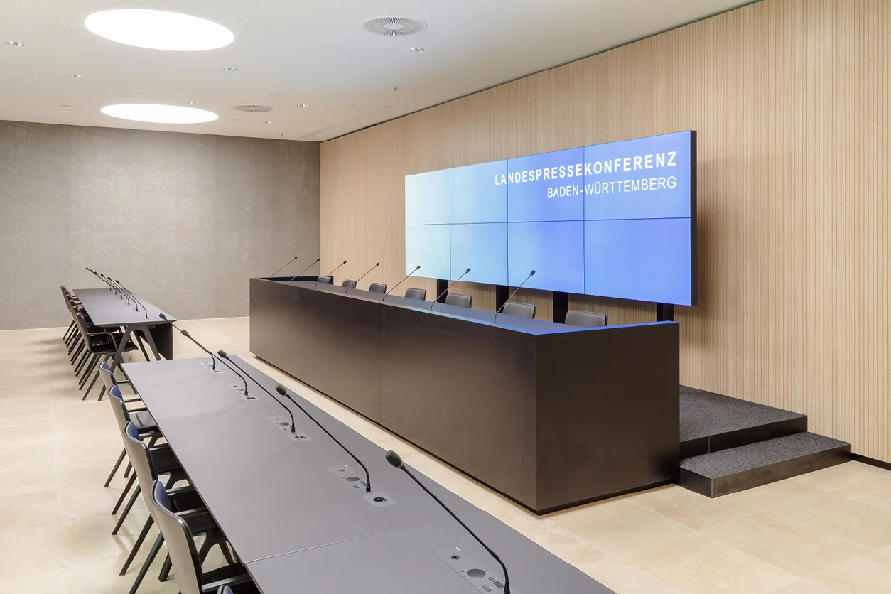 Konferenzraum mit dunkler Podiumstheke, Mikrofontischen und einer hellen, senkrecht gegliederten Holzwandverkleidung.