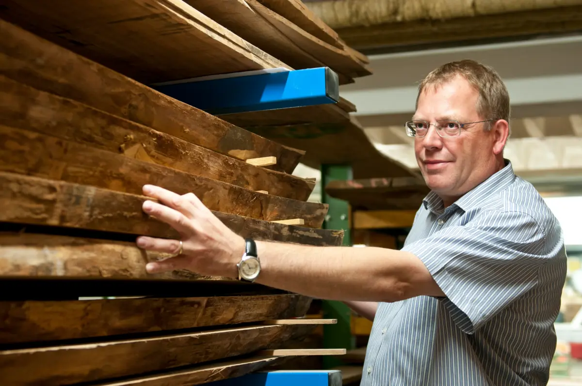 Materialauswahl im Holzlager Ein Mann begutachtet gestapelte Holzbohlen mit natürlichen Kanten und legt seine Hand an eine der Bohlen.
