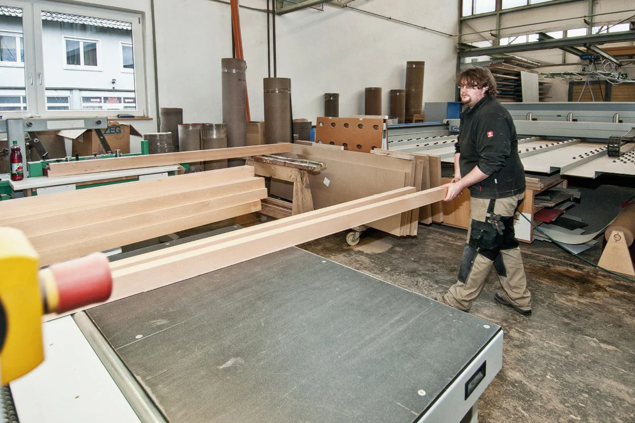 Schleifen und Oberflächentechnik – Werkstückvorbereitung Mitarbeiter hält ein langes Holzwerkstück an einem Maschinentisch in der Schreinerei, daneben liegen weitere Holzteile.